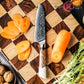 	The Damara Chisai Damascus paring knife resting on a cutting board with a sliced carrot, walnuts, and fresh greens.